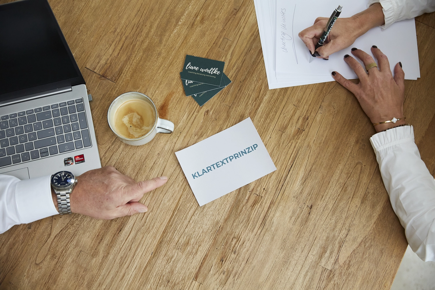 Liane Wodtke Klarheit - Unternehmensberatung und Führungskräfteentwicklung - Draufsicht auf zwei Personen, die an einem Holztisch mit einem Laptop, Kaffee und einer Karte mit der Aufschrift „KLARTEXTPRINZIP“ arbeiten.