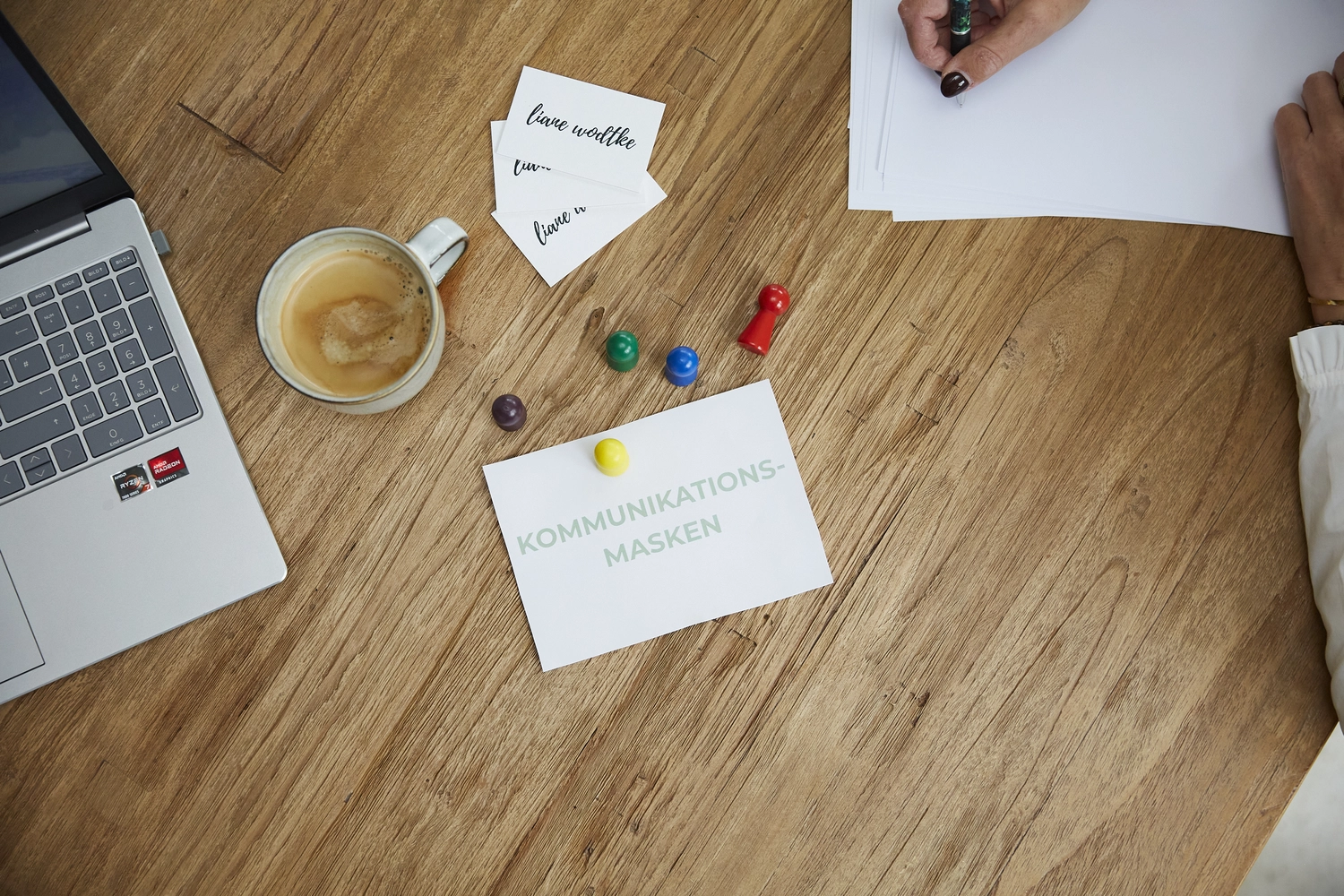 Liane Wodtke Klarheit - Unternehmensberatung und Führungskräfteentwicklung - Eine Draufsicht auf einen Holztisch, an dem jemand auf einem Blatt Papier schreibt, daneben ein Laptop, eine Tasse Kaffee und Notizzettel, darunter einer mit der Aufschrift „KOMMUNIKATIONS-MASKEN“.