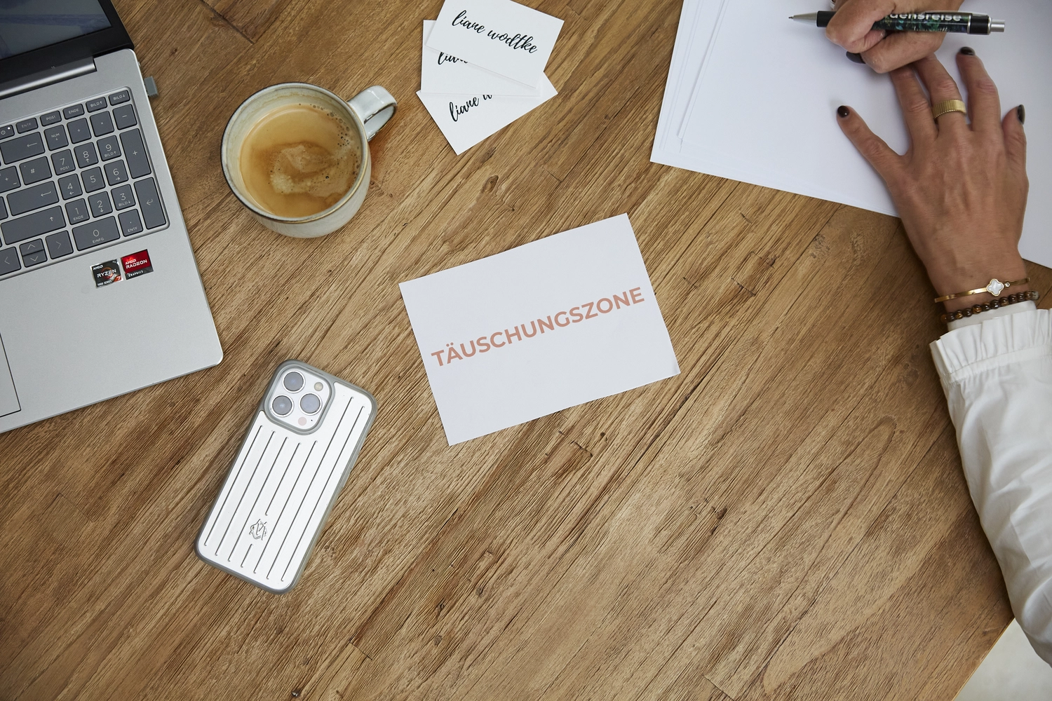 Liane Wodtke Klarheit - Unternehmensberatung und Führungskräfteentwicklung - Draufsicht auf einen Holzschreibtisch mit Laptop, Kaffeetasse, kleinen Visitenkarten und einer weißen Karte mit dem in rosa Schrift gedruckten deutschen Wort „TÄUSCHUNGSZONE“. Rechts ist eine Hand mit Goldschmuck zu sehen, die einen Stift über einem Blatt Papier hält.