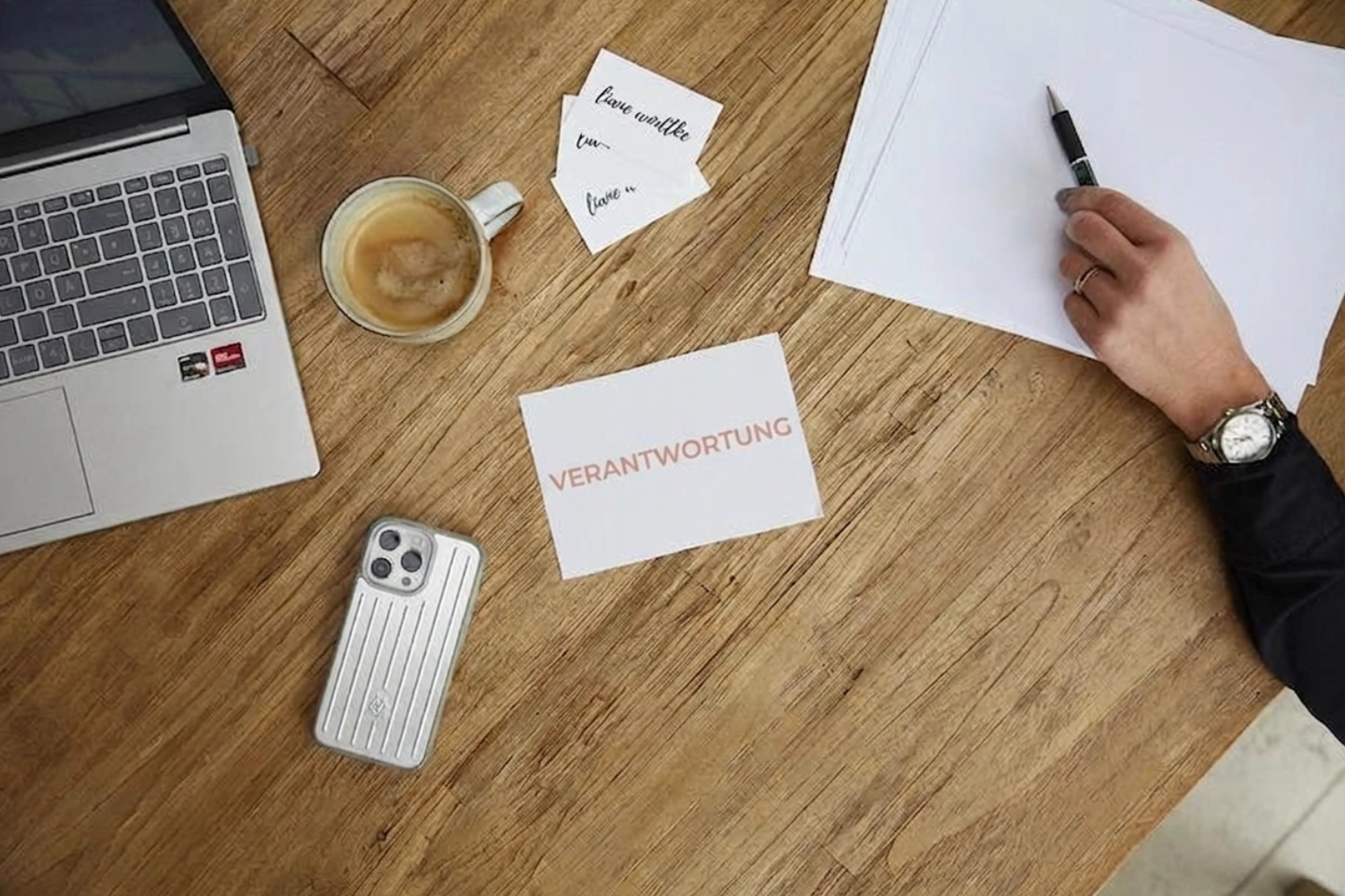 Liane Wodtke Klarheit - Unternehmensberatung und Führungskräfteentwicklung - Eine Aufnahme aus der Vogelperspektive zeigt einen Holzschreibtisch mit Laptop, Kaffeetasse, Smartphone und Visitenkarten. Auf einer großen weißen Karte in der Mitte prangt das deutsche Wort „VERANTWORTUNG“ in orangefarbenen Großbuchstaben. In der oberen rechten Ecke hält eine Hand mit Armbanduhr einen Stift über einen leeren Papierstapel.